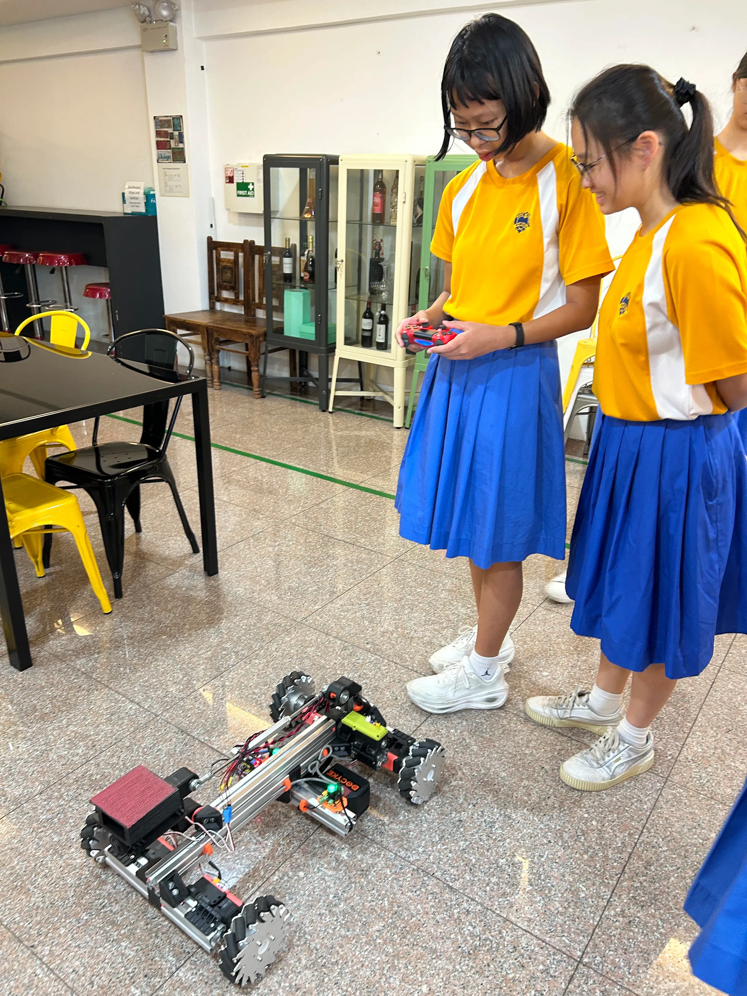 Two students wearing yellow-and-blue school uniforms stand in a learning space, testing a custom-built robotic vehicle on the floor. One student holds a handheld controller while the other observes the robot's performance. The robot, constructed from metal frames, wheels, and electronic components, appears to be part of a STEM or robotics project, providing students with practical experience in design, coding, and problem-solving.
